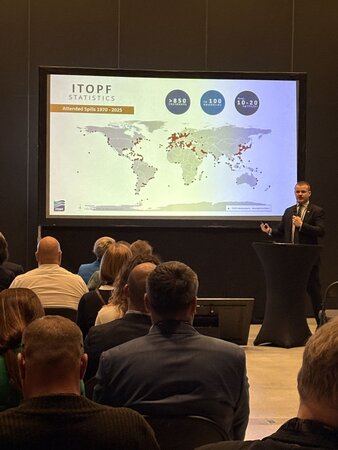 ITOPF presenting during MARISEC 2025 in front of a presentation screen and audience, ITOPF representative holds microphone and is wearing a suit